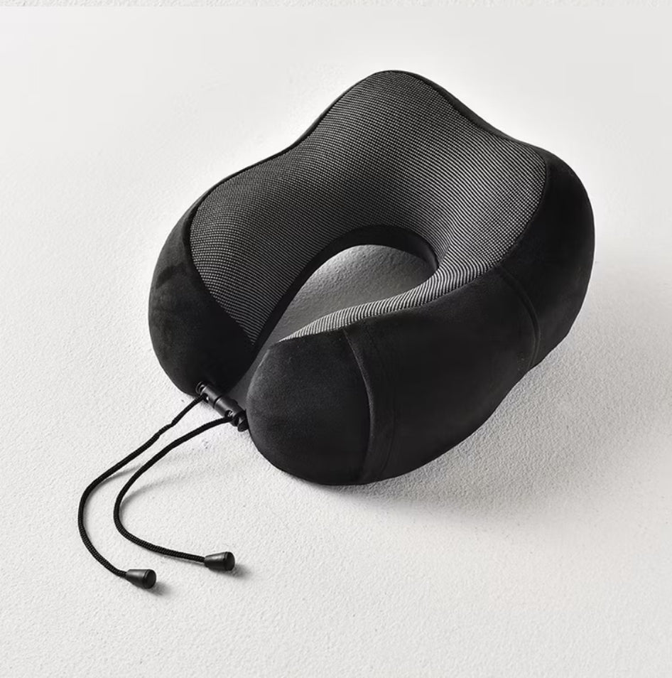 Fotografija prikazuje crni jastuk za vrat sa sivim prugastim detaljima u obliku slova "U". Jastuk leži na beloj površini, a vide se i crne vezice sa plastičnim krajevima.