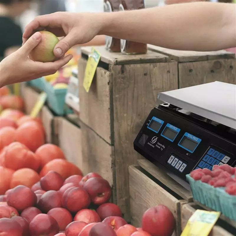 Ruka prodavca predaje jabuku kupcu ispred digitalne vage na pijaci sa vocem.