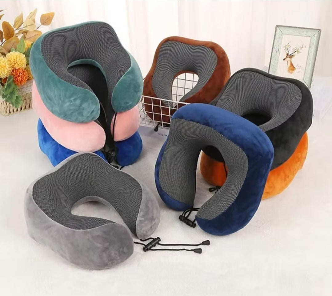 Fotografija prikazuje pet jastuka za vrat različitih boja, uključujući sivu, plavu, roze, zelenu i narandžastu. Jastuci su složeni jedan na drugi na beloj površini, a u pozadini se vide cvetni aranžmani i uramljena slika.