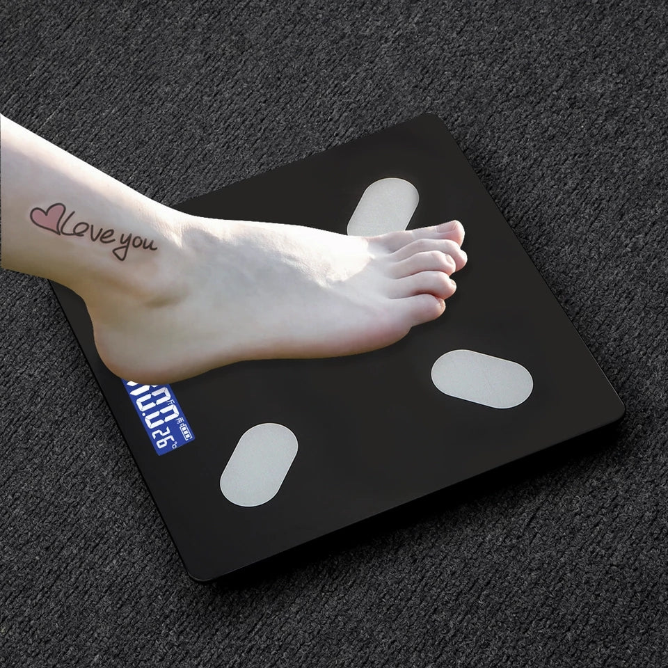 Boso žensko stopalo sa tetovažom "Love you" na gležnju stoji na crnoj pametnoj vagi za telesnu analizu. Digitalni ekran vage prikazuje težinu od 100.26 kg. Vaga ima četiri ovalna senzora od nerđajućeg čelika. Pozadina je tamno siva, moguće tekstilna površina.