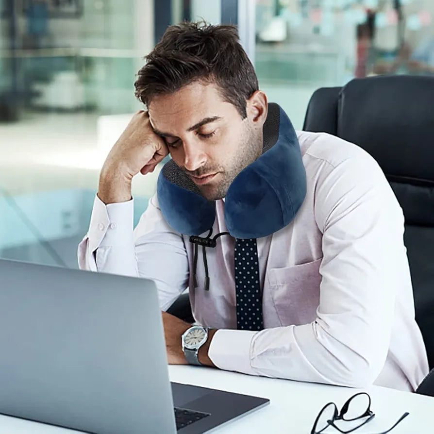 Mladi poslovni čovek sa tamnom kosom i plavim jastukom za vrat, spava na stolu u kancelariji, sa laptopom ispred sebe i naočarima pored. Deluje iscrpljeno i umorno.