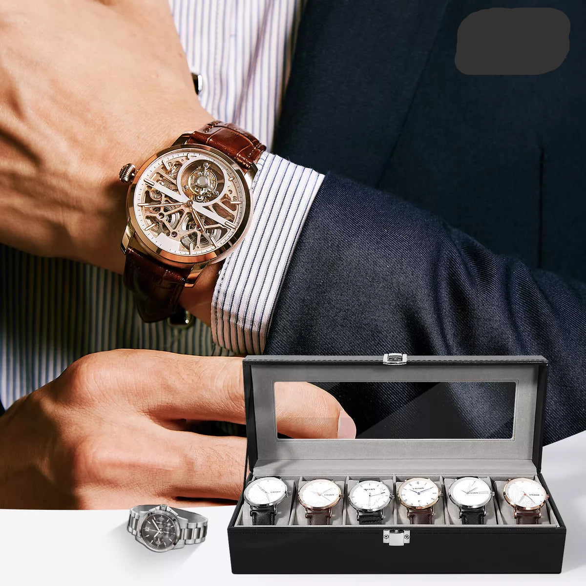 Crna kutija za satove sa šest pregrada i providnim poklopcem. Na ruci se vidi elegantan muški ručni sat sa skeleton mehanizmom i braon kožnom narukvicom. Idealno za čuvanje i prezentaciju muških satova.