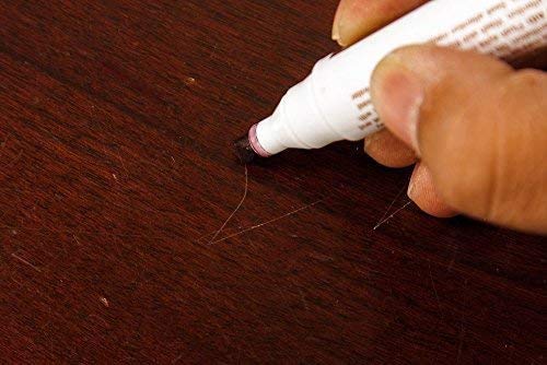Prikaz ruke koja drzi beli marker za popravku drveta i popunjava ogrebotinu na tamno braon drvenoj povrsini. Vrh markera je prislonjen uz ogrebotinu.