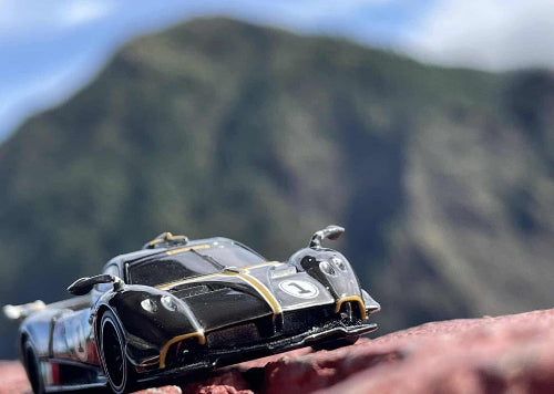 Model crnog Pagani Huayra R automobila sa zlatnim prugama i brojem jedan na haubi postavljen je na neravnoj crvenkastoj povrsini koja podseca na stenu. U pozadini se vidi zamucena slika zelenog brda ili planine ispod svetlog neba sa oblacima.