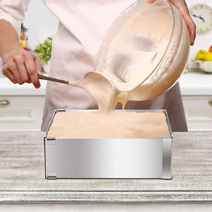 roces izlivanja svetle smese za kolač iz staklene posude u podesivi pravougaoni kalup od nerđajućeg čelika. Kalup se nalazi na svetloj drvenoj površini, a u pozadini se vidi kuhinjski ambijent sa belim elementima. Ruke osobe drže posudu i drvenu špatulu kojom pomažu izlivanje smese.