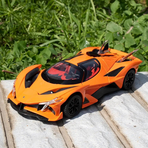Model narandzastog sportskog automobila Apollo Evo sa crnim detaljima i crvenom unutrasnjosti postavljen je na svetloj kamenoj povrsini. U pozadini je zeleno lisce i trava, sto istice jarku narandzastu boju automobila.
