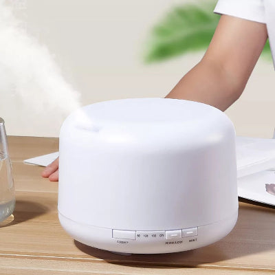 Beli aroma difuzer ovlaživač vazduha na stolu, pored čaše sa vodom i papira, sa maglicom koja izlazi iz vrha.