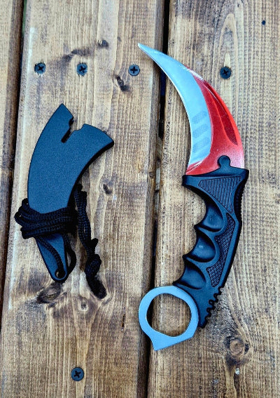 Karambit noz sa crveno-plavim secivom i crnom drskom, replika iz CS:GO igrice. Prikazan je sa futrolom pored. Odlican za kolekciju.