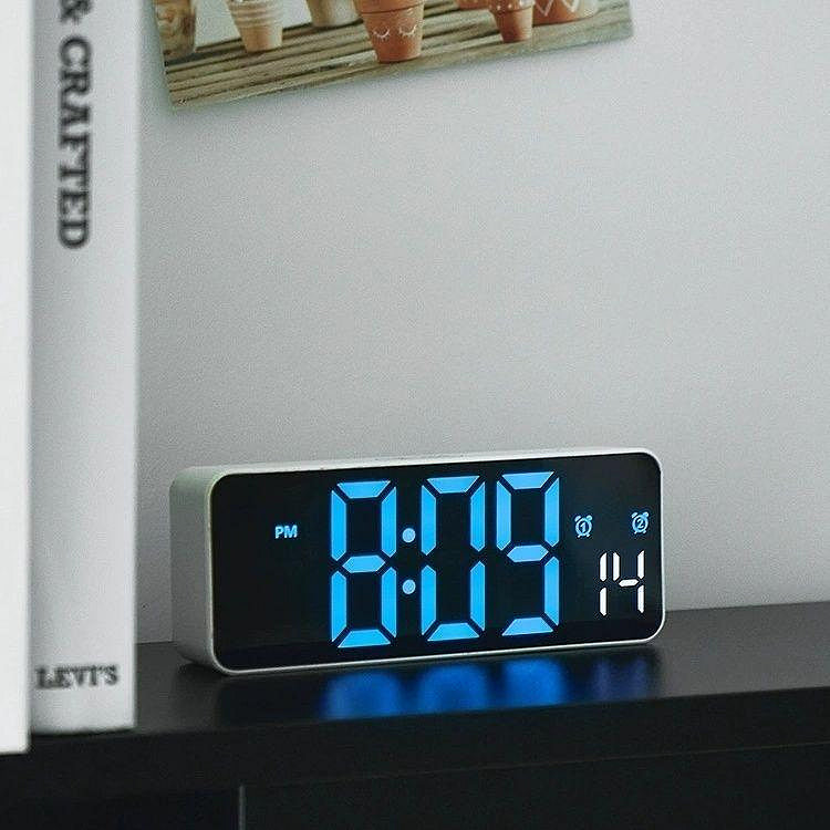 LED digitalni sat sa plavim displejom postavljen na polici pored knjiga