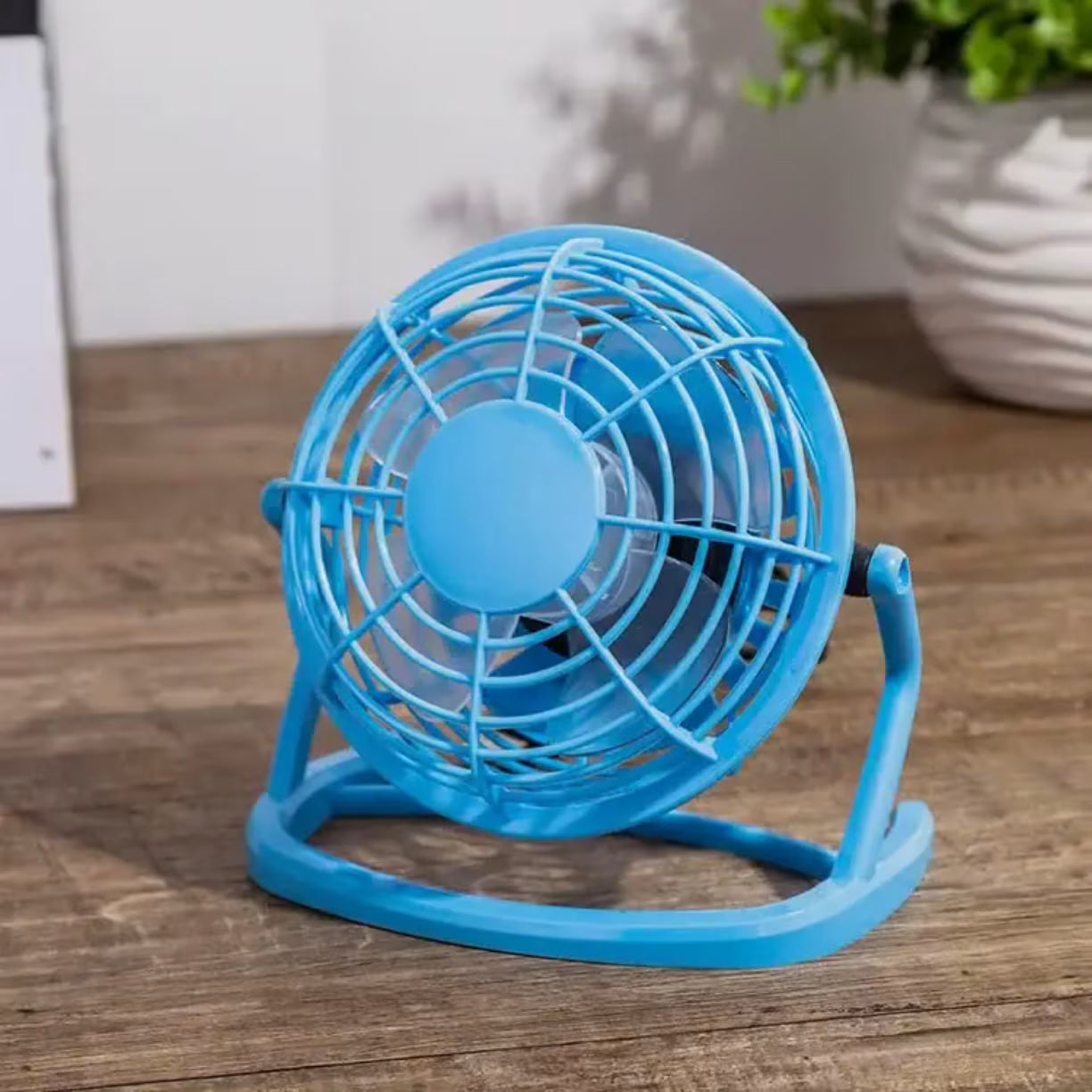 Svetlo plavi mini ventilator sa plasticnim propelerima slikan izbliza na stolu pored saksije