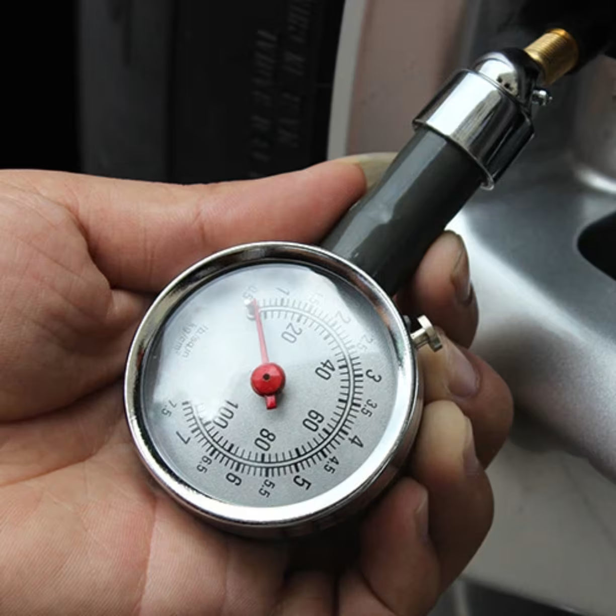 Krupni plan rucnog merenja pritiska gde muska saka drzi sivi analogni uredjaj spojen sa ventilom na automobilu