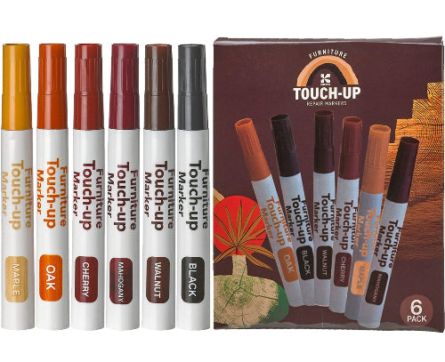 Prikaz seta markera za popravku namestaja u pakovanju. Sa leve strane je osam pojedinacnih markera sa razlicitim bojama cepova i natpisima: zuti (MAPLE), svetlo braon (OAK), crveni (CHERRY), bordo (MAHOGANY), tamno braon (WALNUT) i dva crna (BLACK). Sa desne strane je kartonsko pakovanje sa ilustracijom drvenog goda i sest markera unutra, ciji se cepovi i nazivi boja (OAK, BLACK, WALNUT, CHERRY, MAPLE, MAHOGANY) vide na pakovanju. U donjem desnom uglu pakovanja pise "6 PACK".