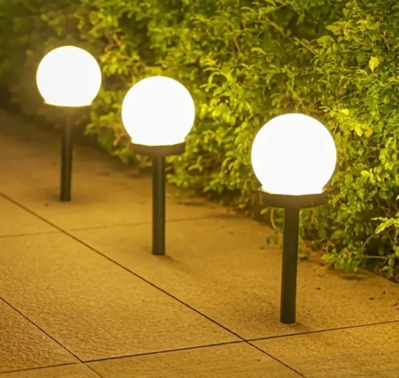 Tri okrugle solarne LED lampe u obliku belih kugli koje svetle toplim svetlom duž popločane staze pored zelenog grmlja