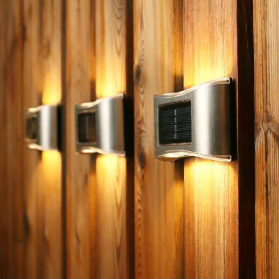 Tri solarne zidne lampe postavljene na drvenoj ogradi. Idealne za osvetljavanje staza i dekoraciju vrta. Bez kablova, napajaju se suncevom energijom.