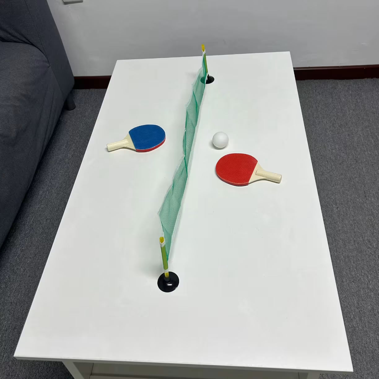 Mini stoni tenis montiran na belom klupskom stolu u dnevnoj sobi sa mrezicom plavim i crvenim reketom